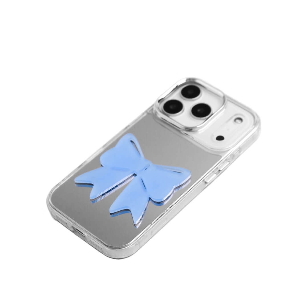 A modern iPhone case featuring a 3D acrylic ribbon ornament with long, flowing tails. Available in Sakura Pink and Azure Blue finishes. The case is shown on both a Reflective Silver metallic base and a Clear Transparent base, showcasing a minimalist yet playful fashion aesthetic.