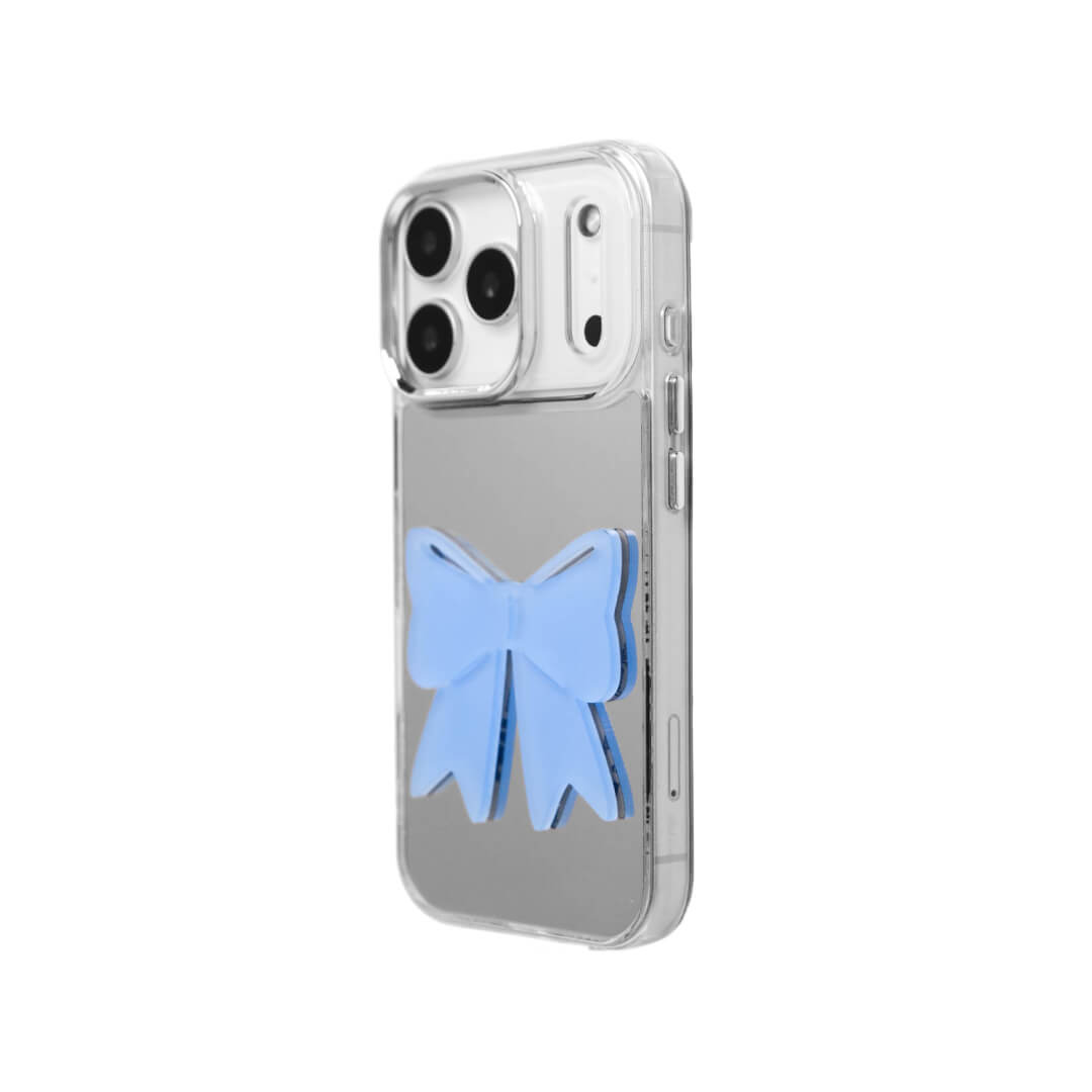 A modern iPhone case featuring a 3D acrylic ribbon ornament with long, flowing tails. Available in Sakura Pink and Azure Blue finishes. The case is shown on both a Reflective Silver metallic base and a Clear Transparent base, showcasing a minimalist yet playful fashion aesthetic.