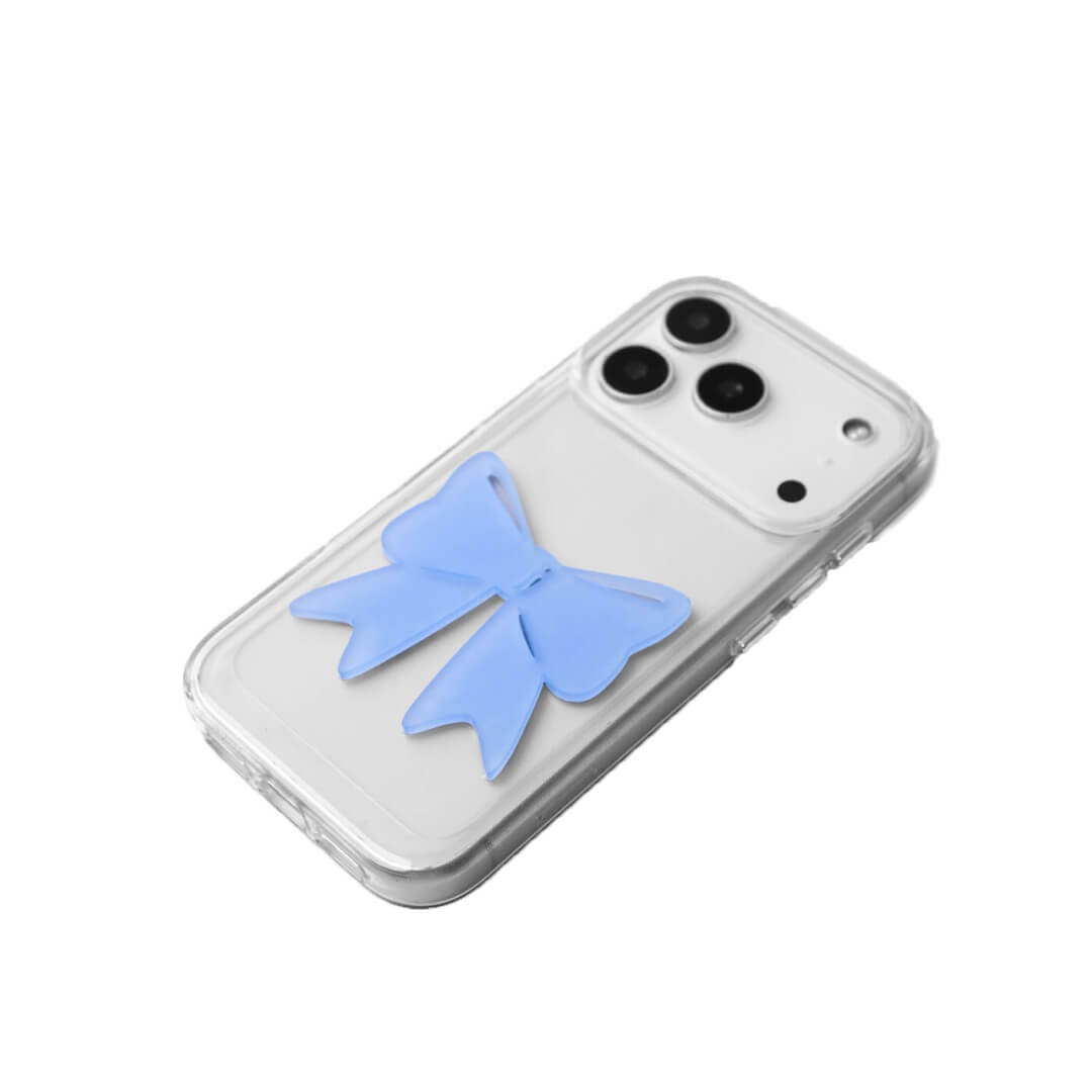 A modern iPhone case featuring a 3D acrylic ribbon ornament with long, flowing tails. Available in Sakura Pink and Azure Blue finishes. The case is shown on both a Reflective Silver metallic base and a Clear Transparent base, showcasing a minimalist yet playful fashion aesthetic.