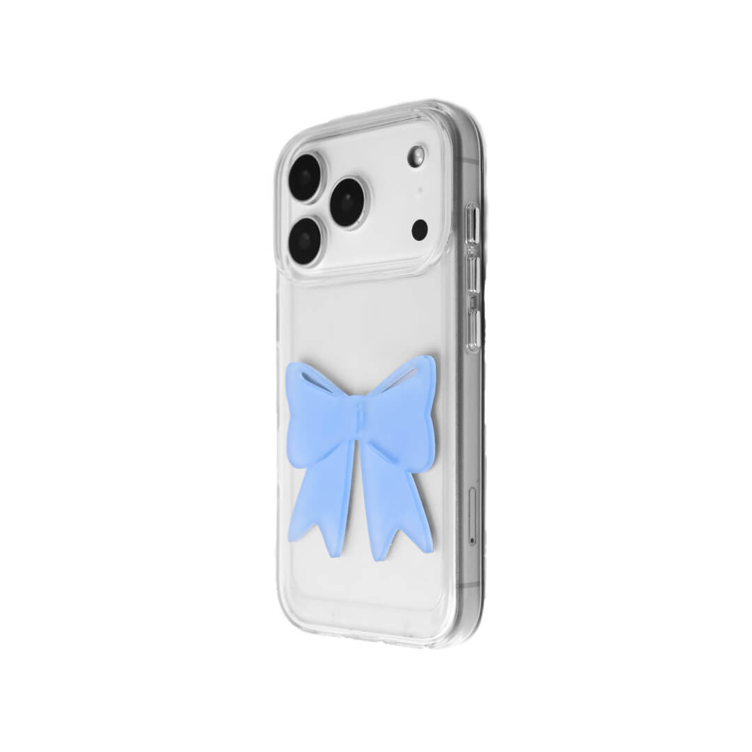 A modern iPhone case featuring a 3D acrylic ribbon ornament with long, flowing tails. Available in Sakura Pink and Azure Blue finishes. The case is shown on both a Reflective Silver metallic base and a Clear Transparent base, showcasing a minimalist yet playful fashion aesthetic.