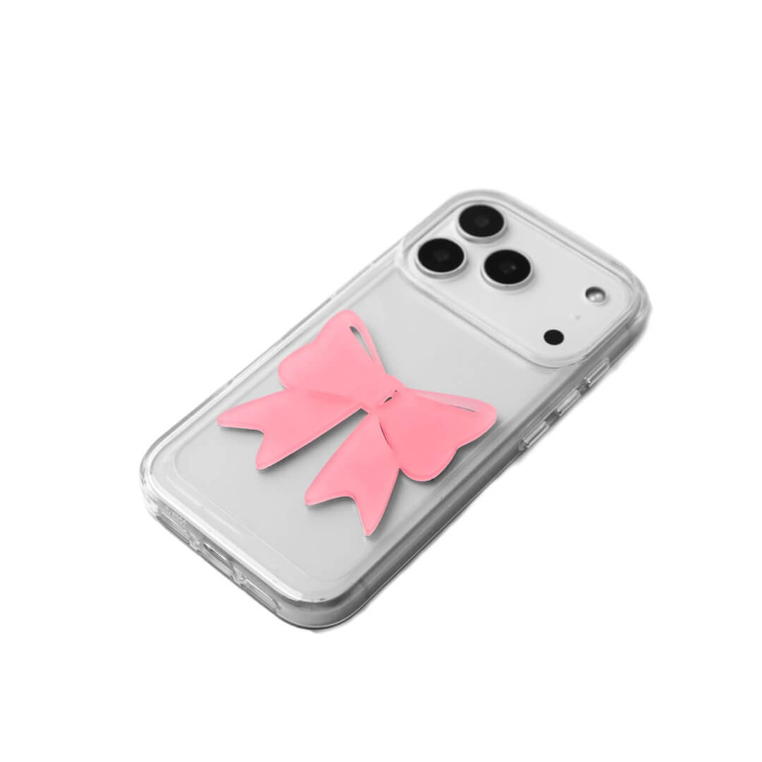 A modern iPhone case featuring a 3D acrylic ribbon ornament with long, flowing tails. Available in Sakura Pink and Azure Blue finishes. The case is shown on both a Reflective Silver metallic base and a Clear Transparent base, showcasing a minimalist yet playful fashion aesthetic.