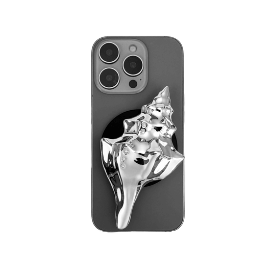 Glossy silver, white and yellow conch shell-shaped phone grip with sculptural ridges and rhinestone accents, mounted on a black base and attached to a gray iPhone case.