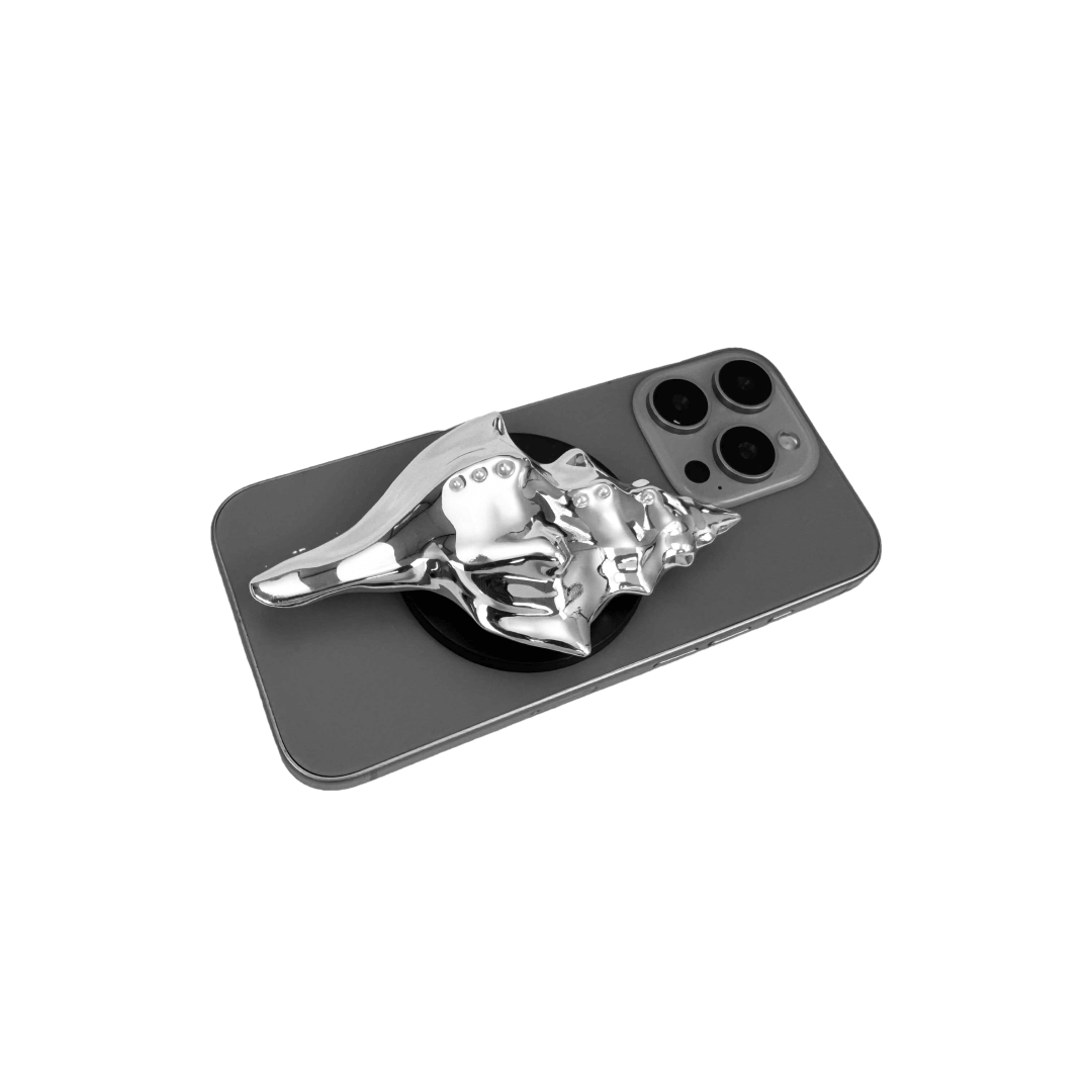 Glossy silver, white and yellow conch shell-shaped phone grip with sculptural ridges and rhinestone accents, mounted on a black base and attached to a gray iPhone case.