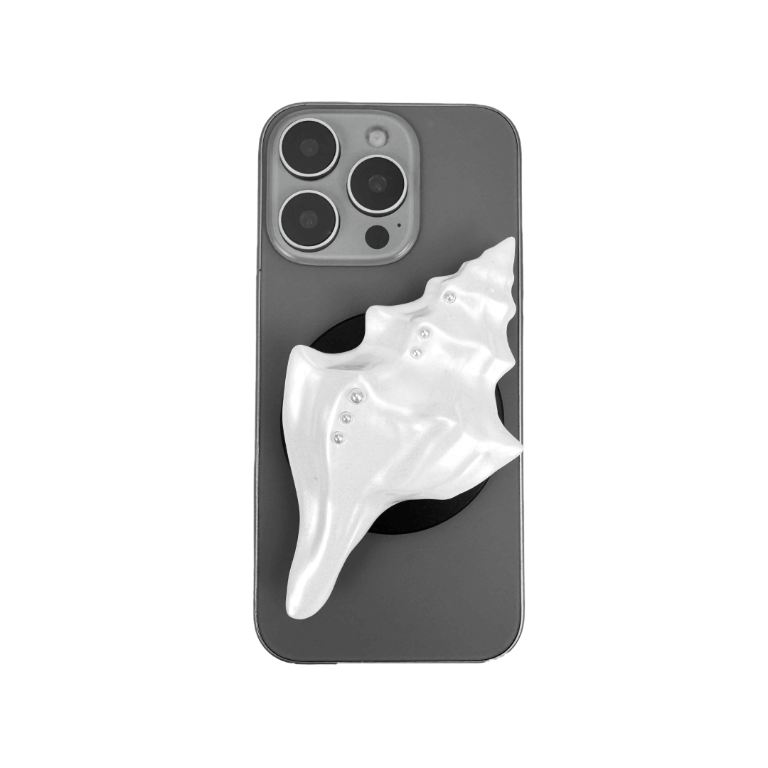 Glossy silver, white and yellow conch shell-shaped phone grip with sculptural ridges and rhinestone accents, mounted on a black base and attached to a gray iPhone case.