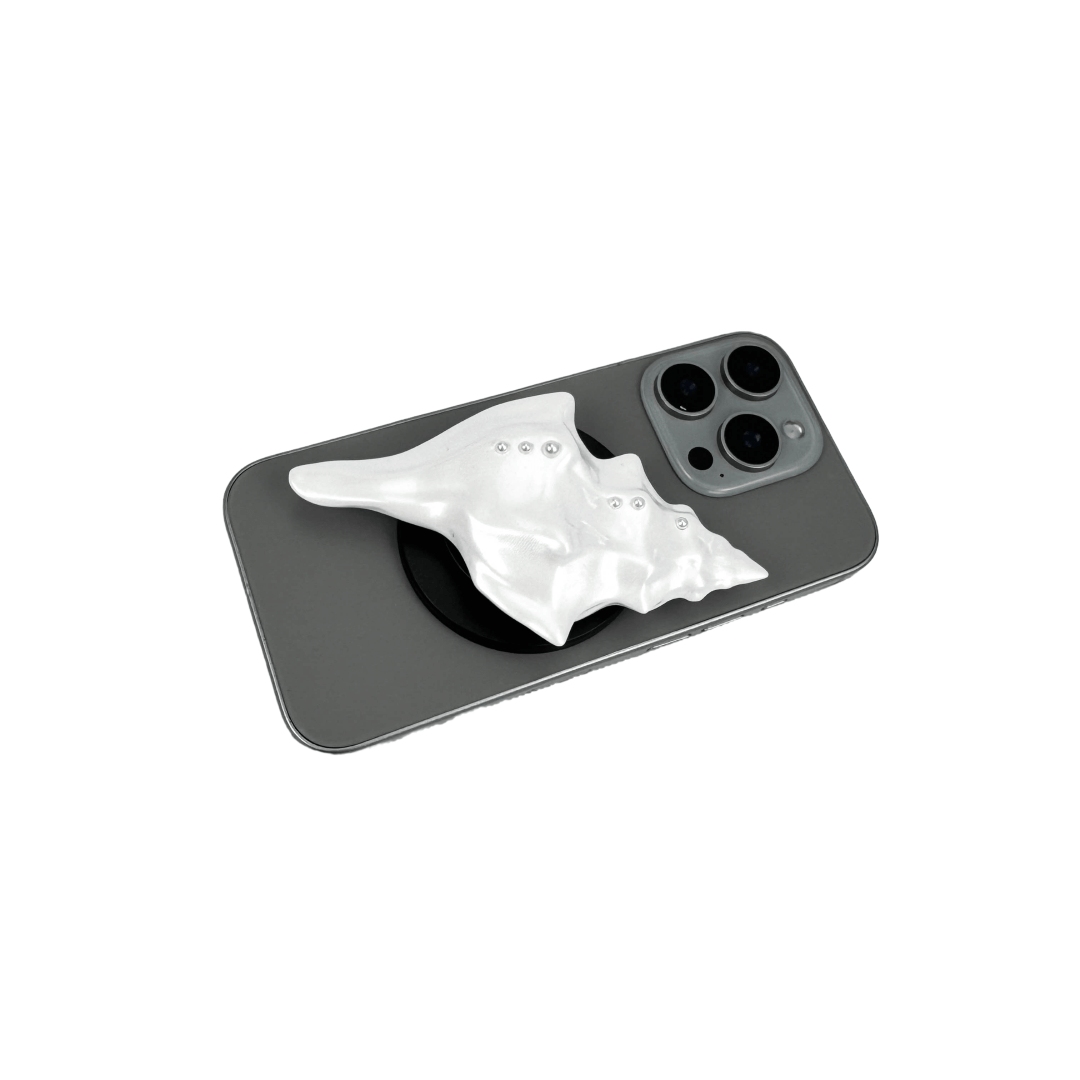 Glossy silver, white and yellow conch shell-shaped phone grip with sculptural ridges and rhinestone accents, mounted on a black base and attached to a gray iPhone case.