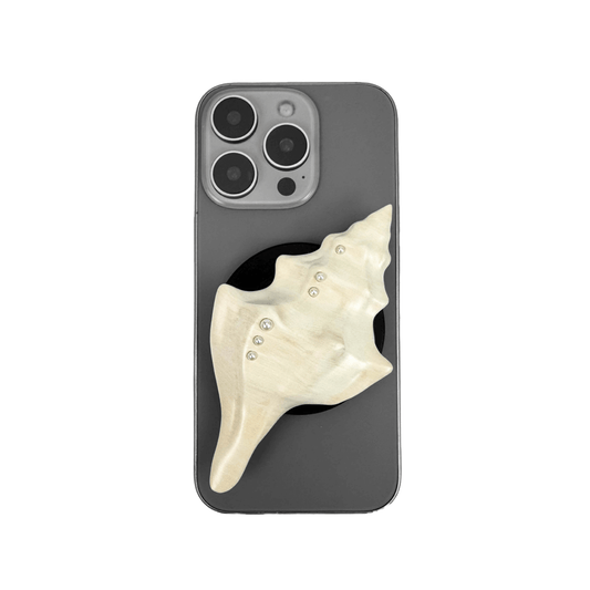 Glossy silver, white and yellow conch shell-shaped phone grip with sculptural ridges and rhinestone accents, mounted on a black base and attached to a gray iPhone case.