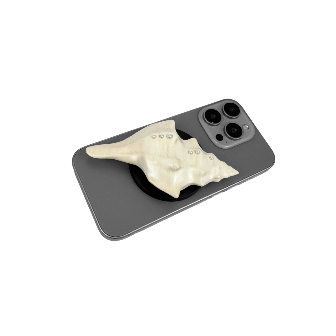 Glossy silver, white and yellow conch shell-shaped phone grip with sculptural ridges and rhinestone accents, mounted on a black base and attached to a gray iPhone case.