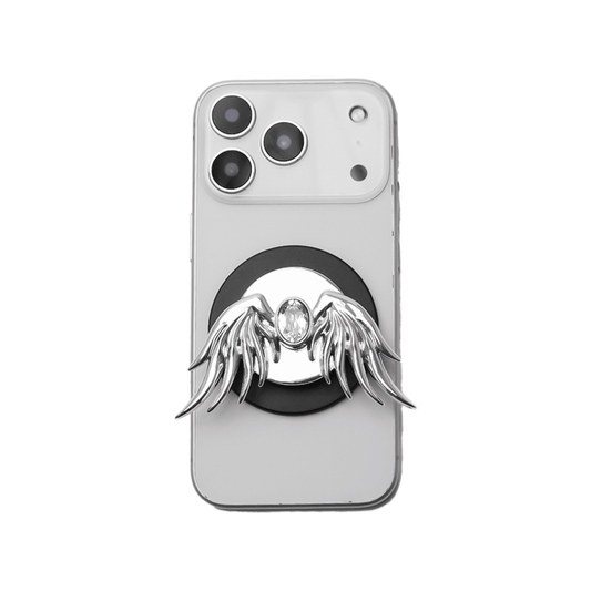The Gem Angel Wings phone grip featuring intricately sculpted 3D wings with a sparkling central Swarovski crystal. Shown in both the reflective Silver Chrome and pure White finish options, attached to the back of a smartphone.