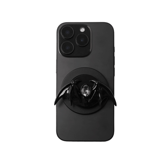 A 3D sculpted demon wing phone grip featuring a central black Swarovski crystal. The product is shown in two finish options: a reflective Silver Chrome that contrasts with the dark gem, and a Sleek Black finish for a full monochromatic look. It is designed to function as both a secure hand grip and a desktop stand.