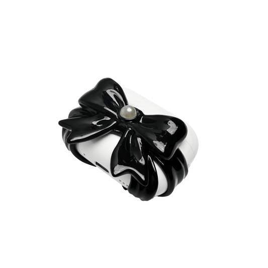 A pair of protective covers for Apple AirPods (or AirPods Pro) charging case. Each cover is white and features a large, 3D sculpted bow charm centered on the front. One bow is glossy black and the other is pure white, with a single white pearl embedded in the center of each.