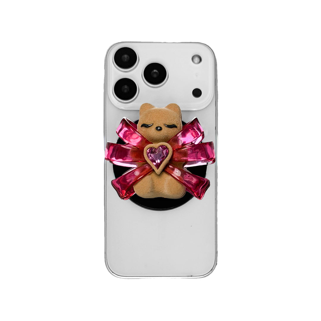 Ribbon Magic Bear Phone Grip in Pink Edition featuring a glossy pink ribbon with a beige gummy bear at the center, holding a sparkling pink heart-shaped crystal.