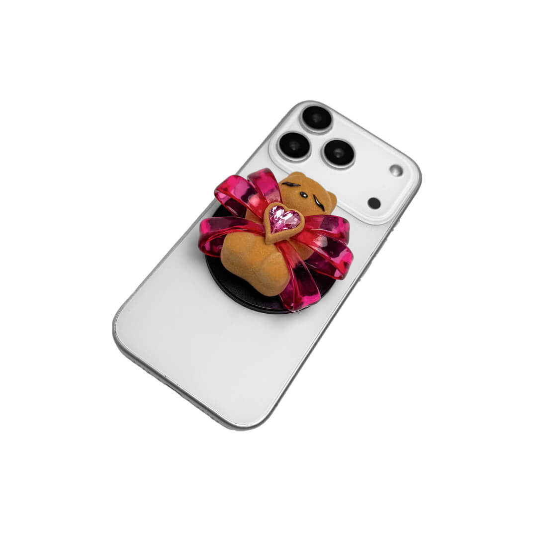 Ribbon Magic Bear Phone Grip in Pink Edition featuring a glossy pink ribbon with a beige gummy bear at the center, holding a sparkling pink heart-shaped crystal.