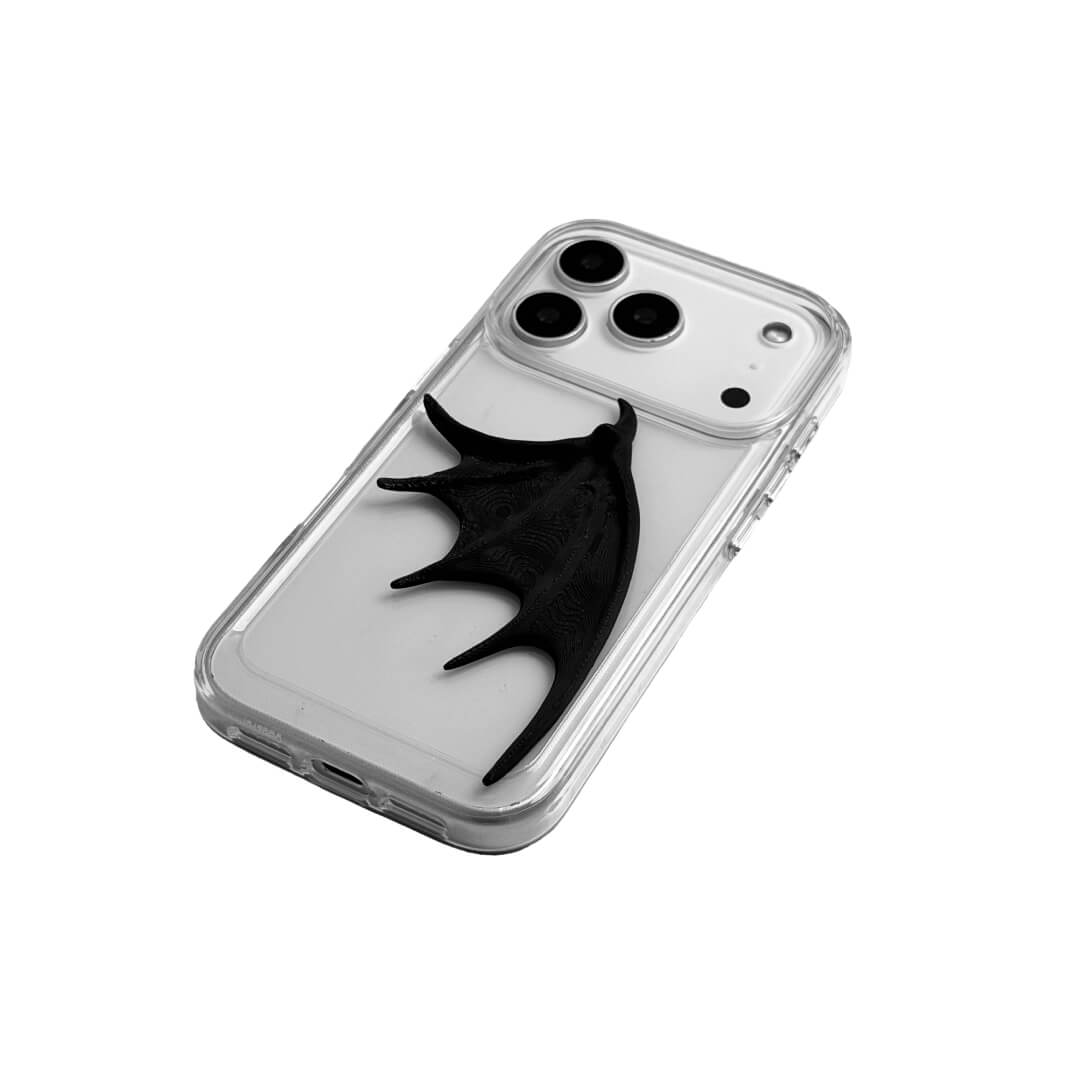 A high-fashion iPhone case featuring a large, 3D-printed single demon wing with a natural matte black finish. The design is presented in two styles: an asymmetric wing on a Clear Transparent base, and the same sculptural wing on a Black Leather textured base, showcasing a moody and avant-garde aesthetic.