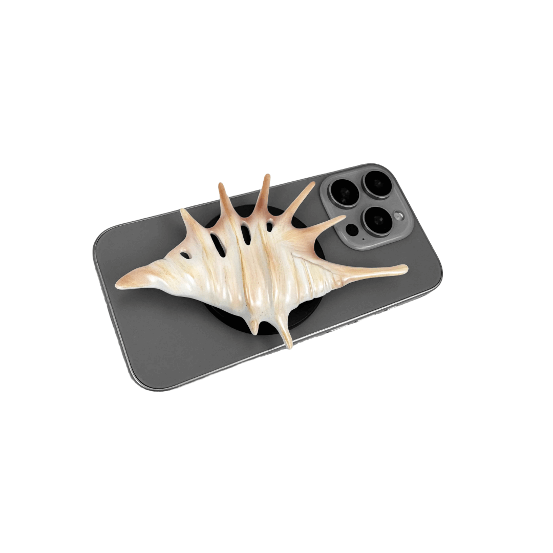 Matte silver, white and yellow spiked conch shell phone grip with sculptural spikes and spiral ridges, mounted on a black base and attached to a gray iPhone case.