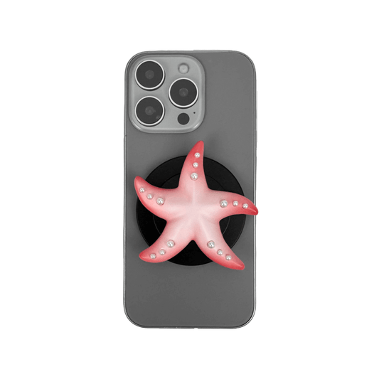 Glossy white, silver, gold and pink starfish-shaped phone grip with embedded rhinestones, mounted on a black base and attached to a gray iPhone case. 