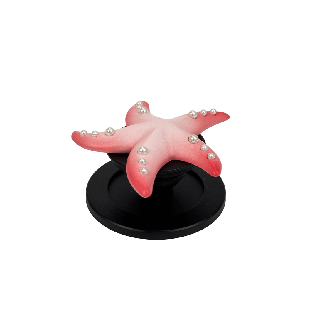 Glossy white, silver, gold and pink starfish-shaped phone grip with embedded rhinestones, mounted on a black base and attached to a gray iPhone case. 
