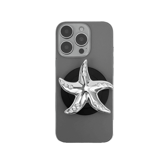 Glossy white, silver, gold and pink starfish-shaped phone grip with embedded rhinestones, mounted on a black base and attached to a gray iPhone case. 