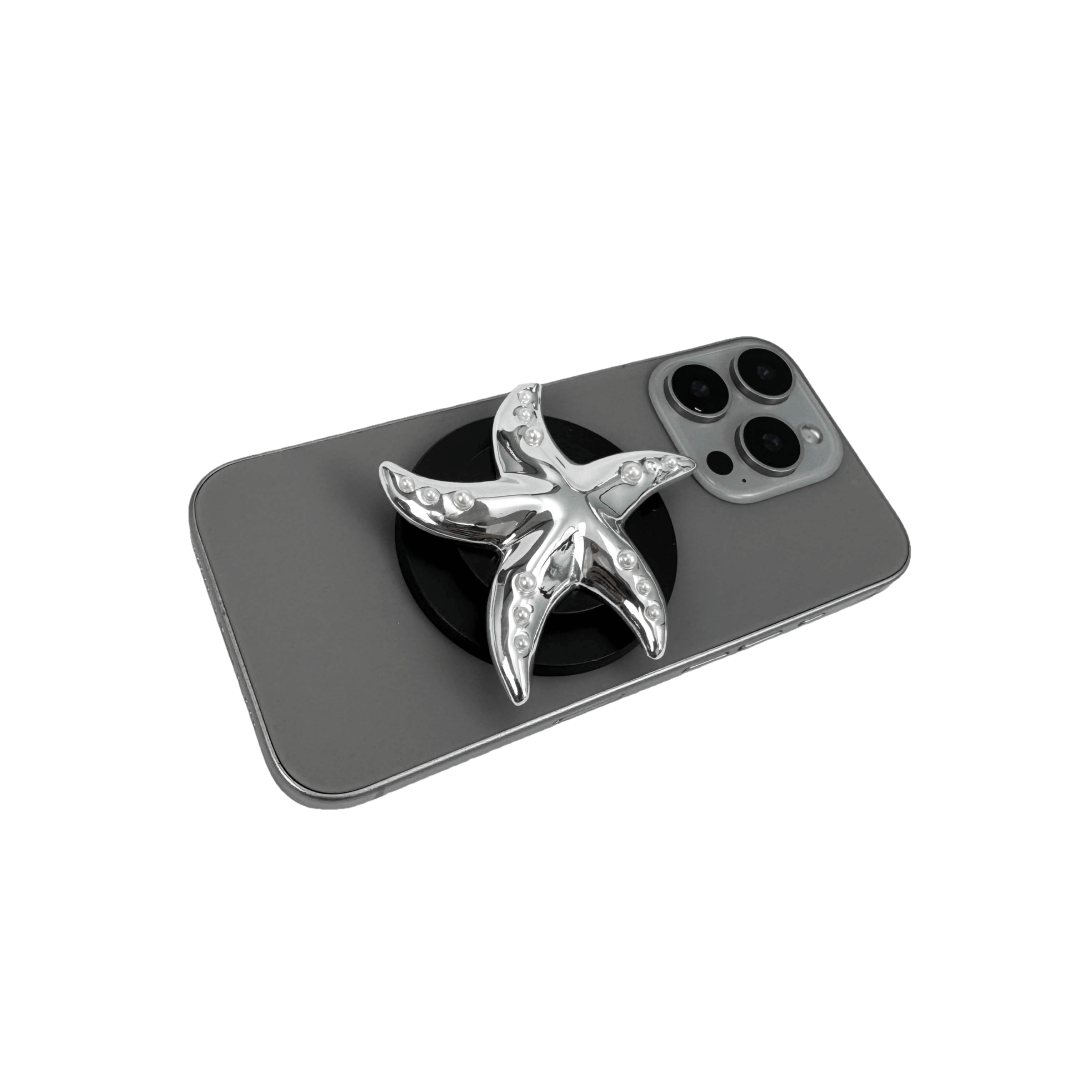 Glossy white, silver, gold and pink starfish-shaped phone grip with embedded rhinestones, mounted on a black base and attached to a gray iPhone case. 