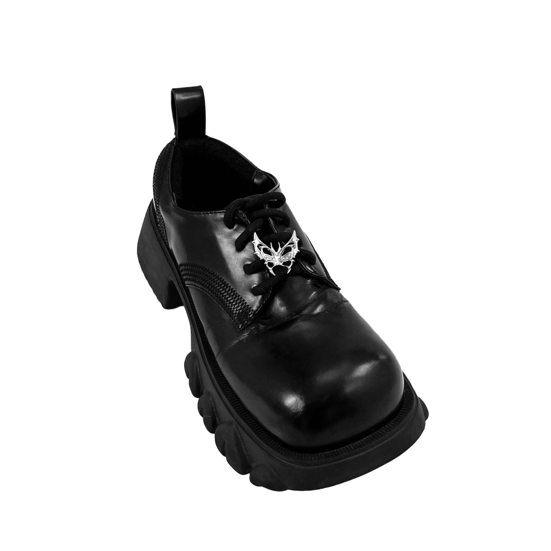 Sculptural chrome wing-shaped shoe accessory clipped onto the side or lace area of a black leather shoe.