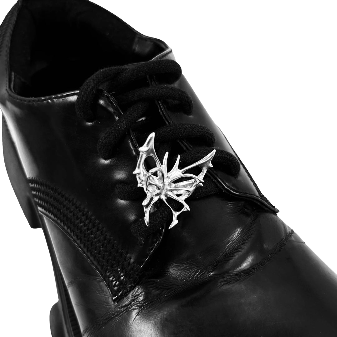 Sculptural chrome wing-shaped shoe accessory clipped onto the side or lace area of a black leather shoe.